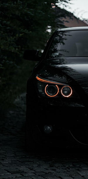 Black BMW M5 E60 Front Headlight Close-Up with Orange Halo Rings on Cobblestone HD phone wallpaper