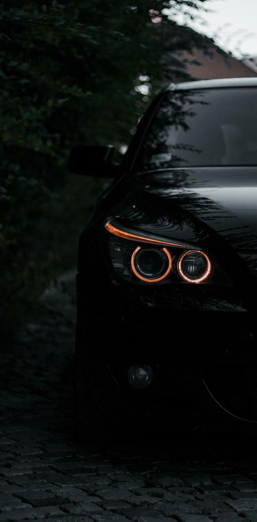 Black BMW M5 E60 Front Headlight Close-Up with Orange Halo Rings on Cobblestone HD phone wallpaper