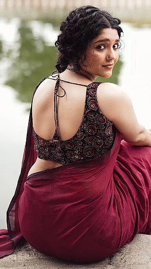 Rithika Singh in a deep maroon saree with an embroidered black blouse, seated by a calm water body HD phone wallpaper