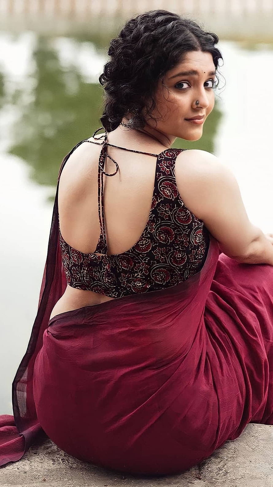 Rithika Singh in a deep maroon saree with an embroidered black blouse, seated by a calm water body HD phone wallpaper
