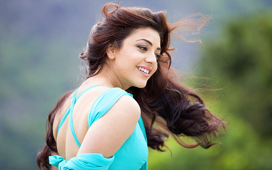 Kajal Aggarwal in a turquoise dress with windblown hair against a soft green outdoor backdrop HD wallpaper