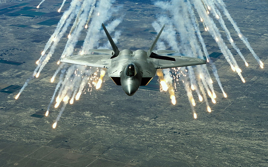 F-22 Raptor fighter jet releasing flares over a patchwork agricultural landscape HD wallpaper