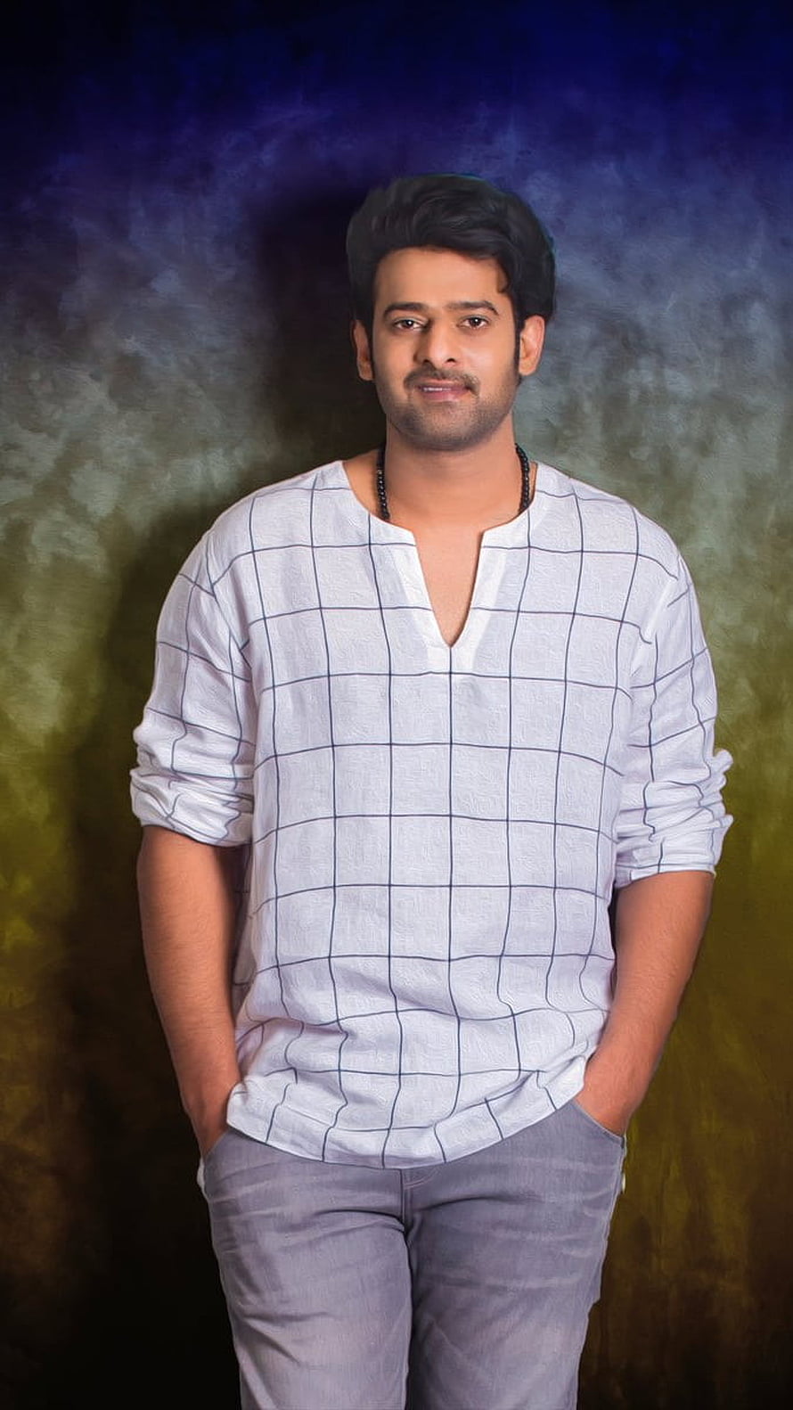 Prabhas in a white checkered shirt and grey trousers against a textured studio backdrop HD phone wallpaper