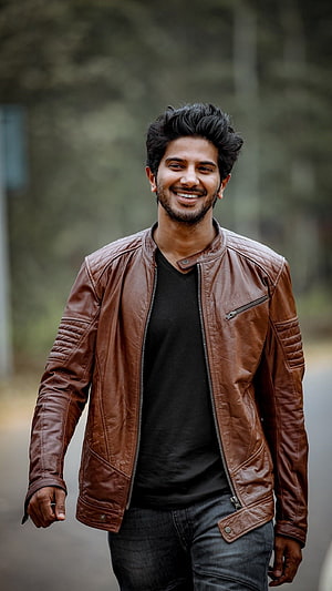 Dulquer Salmaan in a brown leather jacket and black t-shirt smiling outdoors HD phone wallpaper