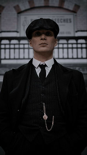 Tommy Shelby in a dark pinstripe suit and flat cap standing before an industrial brick wall HD phone wallpaper