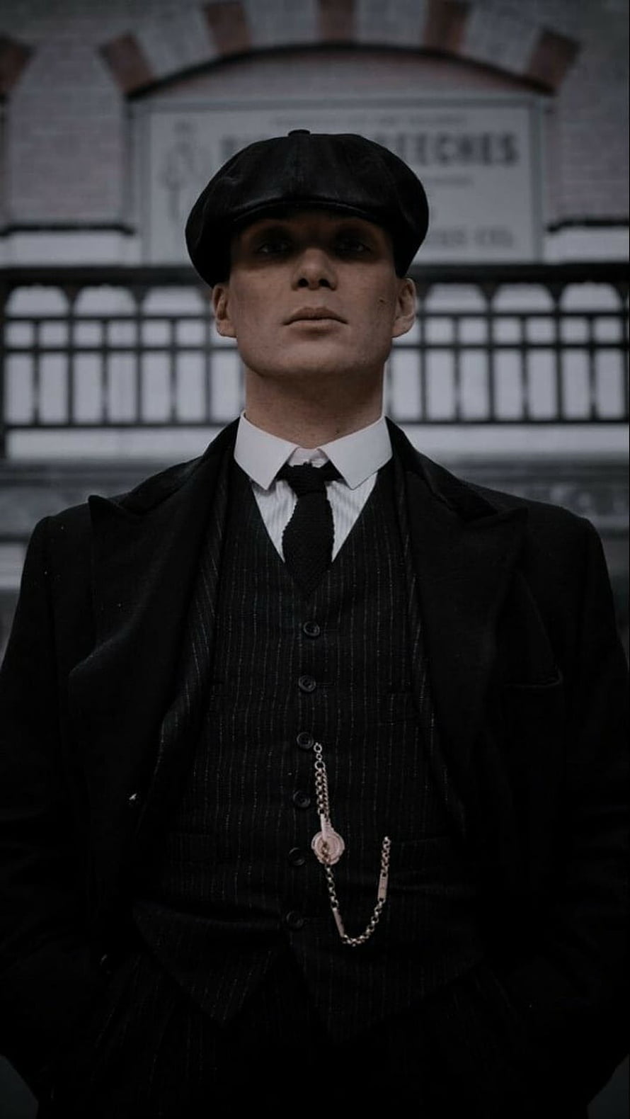 Tommy Shelby in a dark pinstripe suit and flat cap standing before an industrial brick wall HD phone wallpaper