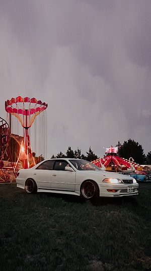 White Toyota Mark II JZX100 Sedan Parked at a Nighttime Carnival with Neon Rides HD phone wallpaper