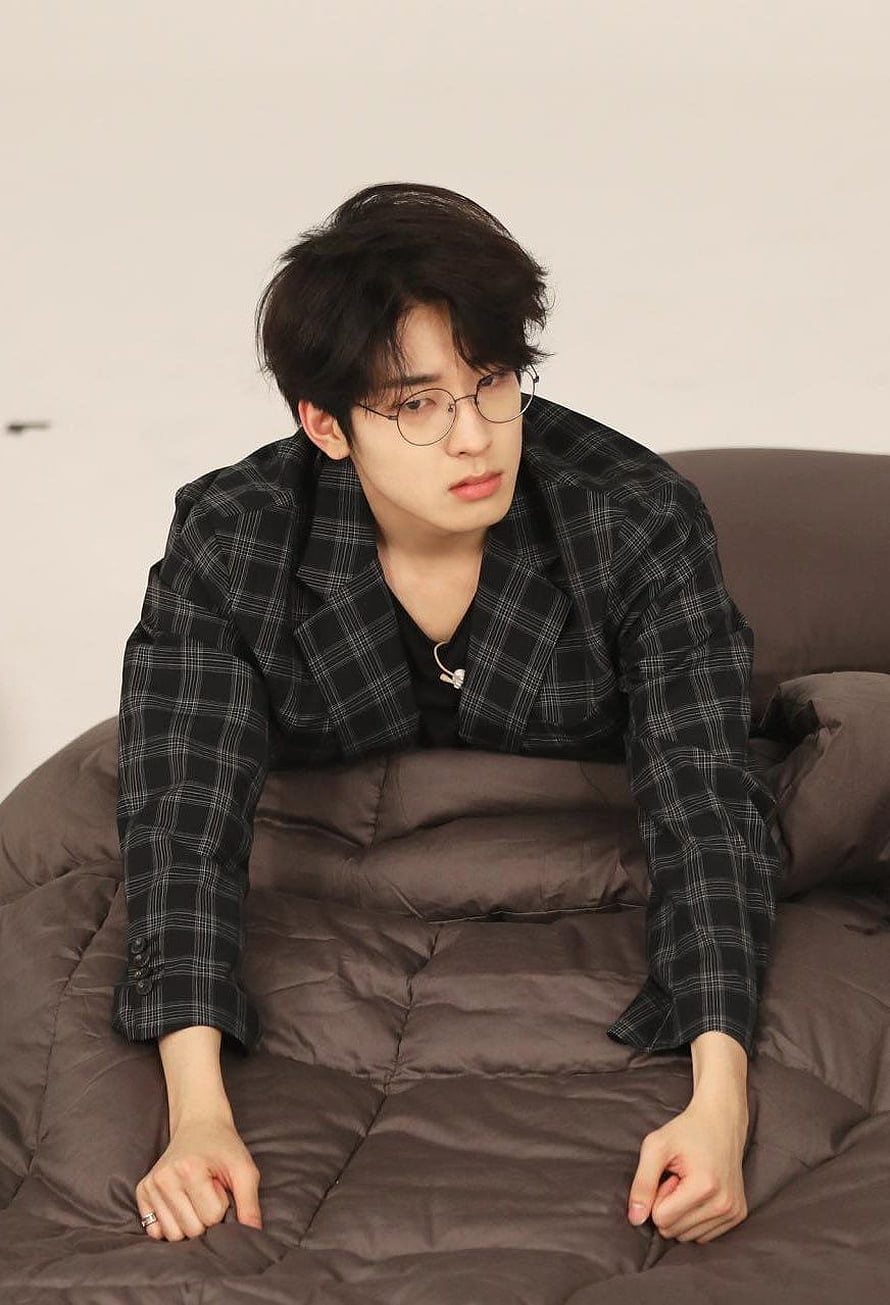 Jeon Wonwoo in a black plaid blazer and round glasses sitting on a brown bed HD phone wallpaper