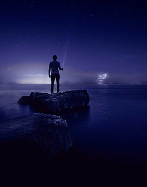 Silhouette of a person standing on a rock gazing at a starry night sky over calm water HD phone wallpaper