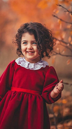 Smiling Curly-Haired Girl in Red Dress with Autumn Foliage Background HD phone wallpaper