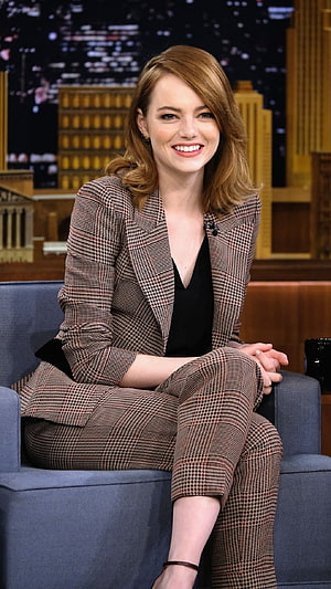 Emma Stone in a houndstooth suit seated on a talk show set with a city skyline backdrop HD phone wallpaper
