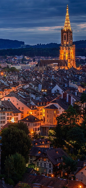 Bern Switzerland Cityscape at Dusk with Illuminated Cathedral Spire and Historic Roofs HD phone wallpaper