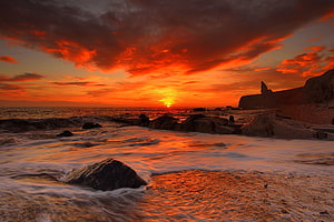 Vibrant Orange Sunset Over Rocky Shoreline With Dramatic Clouds And Gentle Waves HD wallpaper