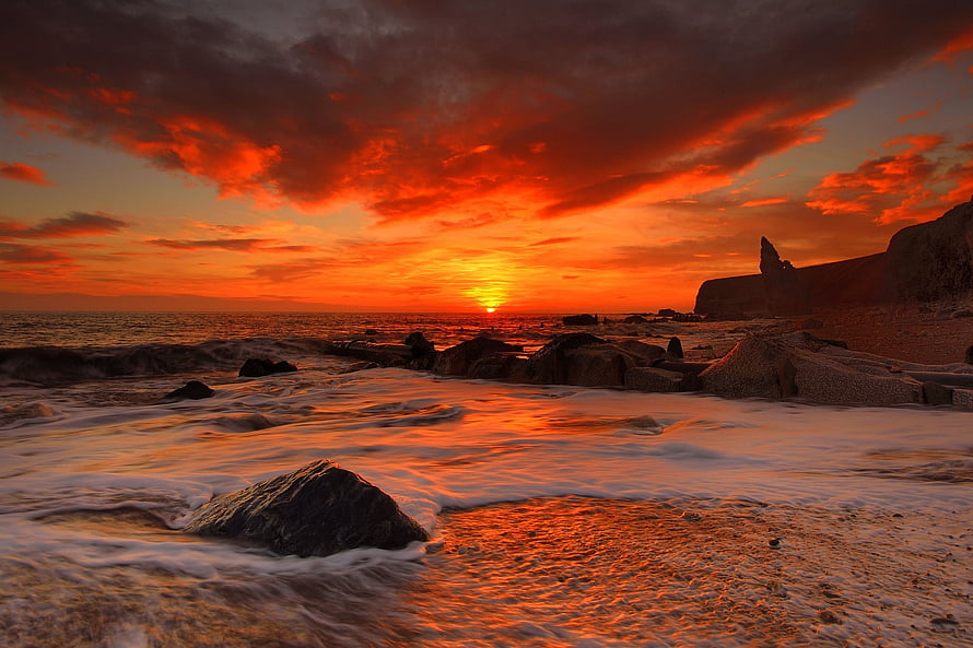 Vibrant Orange Sunset Over Rocky Shoreline With Dramatic Clouds And Gentle Waves HD wallpaper