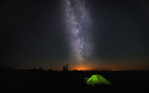 Glowing green tent beneath the Milky Way with mountain silhouettes and a warm horizon glow HD wallpaper
