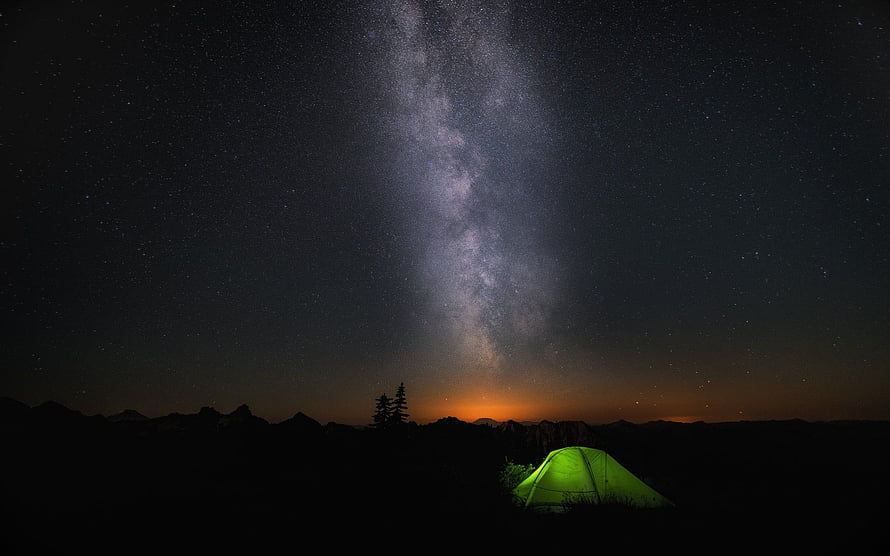 Glowing green tent beneath the Milky Way with mountain silhouettes and a warm horizon glow HD wallpaper