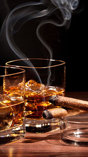 Two glasses of amber whiskey with ice and two lit cigars resting on a wooden table against a dark background HD phone wallpaper