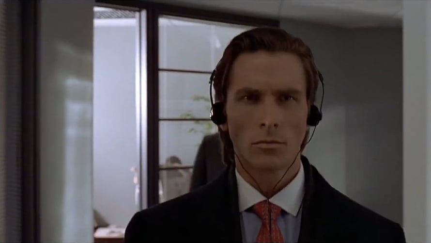 Patrick Bateman in a dark suit and red tie wearing a black headset in a modern office HD wallpaper