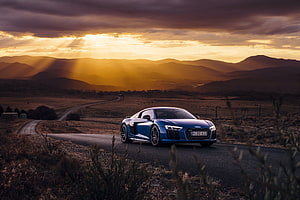 Blue Audi R8 V10 on a winding country road at golden hour with dramatic mountain sunset HD wallpaper