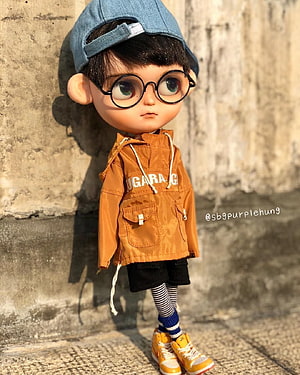 Stylish BJD Doll Boy Wearing Round Glasses, Denim Cap, and Orange Jacket Against Concrete Wall HD phone wallpaper
