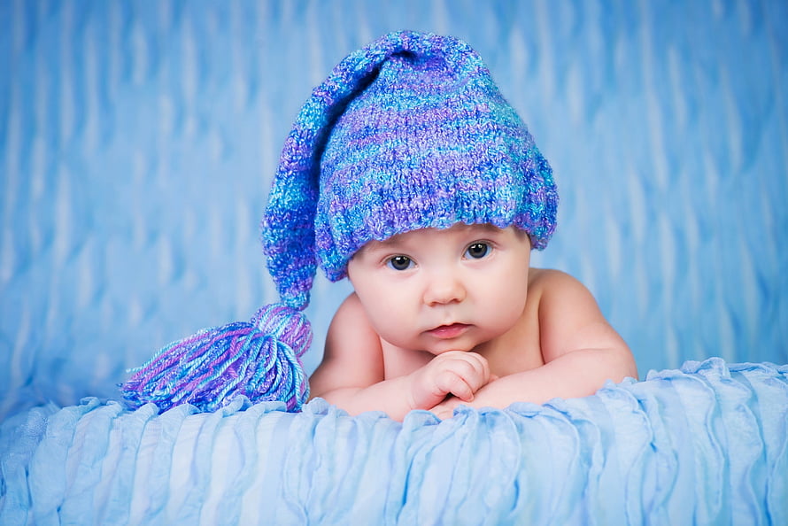 Adorable baby boy wearing a colorful knitted winter hat with a tassel against a soft blue backdrop HD wallpaper
