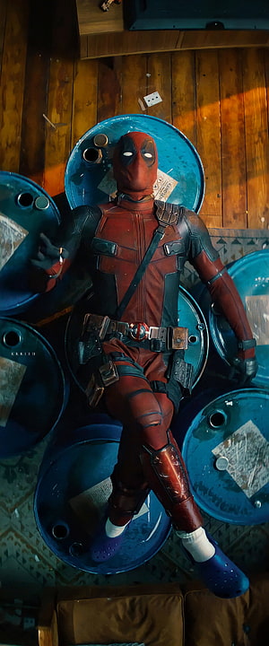 Deadpool in red and black suit reclining on blue barrels in dimly lit room HD phone wallpaper