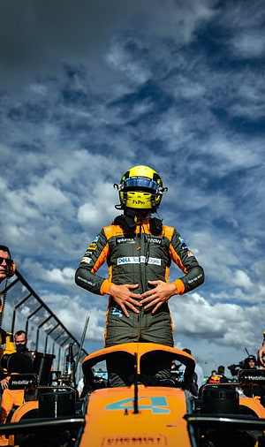 Lando Norris standing on his McLaren Formula 1 car under a dramatic cloudy sky HD phone wallpaper