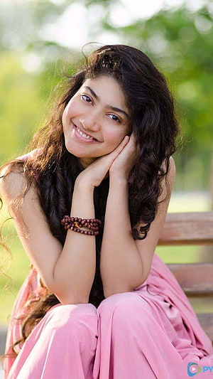 Sai Pallavi smiling portrait in a pink dress sitting on a wooden bench outdoors HD phone wallpaper