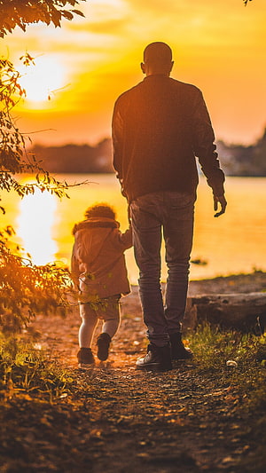 Father and child walking together along a lakeside path at golden hour sunset HD phone wallpaper