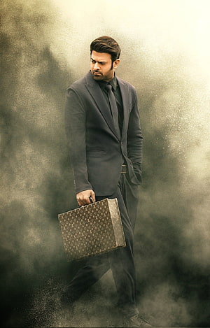 Prabhas in a dark grey suit holding a Louis Vuitton briefcase against a smoky backdrop HD phone wallpaper