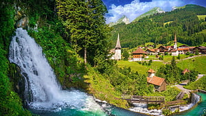 Jaunbach Waterfall cascading into a turquoise river beside a Swiss alpine village with church spires and green meadows HD wallpaper