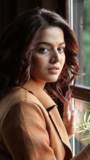 Wamiqa Gabbi in a warm brown coat gazing thoughtfully from a window with soft natural light HD phone wallpaper