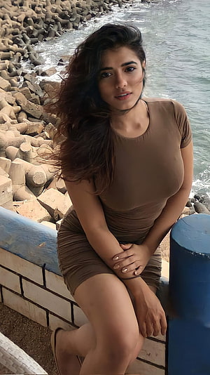 Kethika Sharma Sitting on a Seaside Wall with Ocean and Breakwater Rocks Behind Her HD phone wallpaper