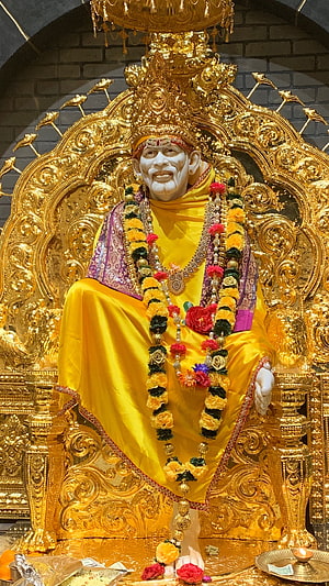 Sai Baba seated on ornate golden throne wearing yellow robes and floral garlands in temple setting HD phone wallpaper