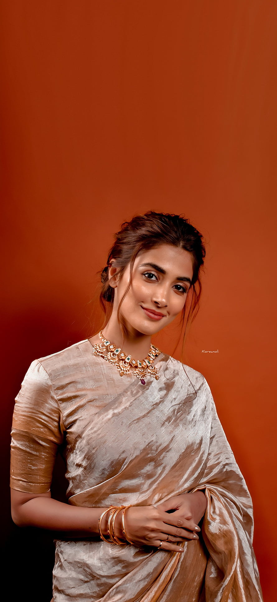 Pooja Hegde in a shimmering beige silk saree with ornate gold jewelry against a warm terracotta backdrop HD phone wallpaper