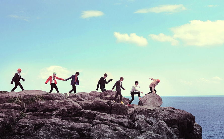 BTS Members Walking Along Rocky Coastal Cliffs Under a Bright Blue Sky HD wallpaper