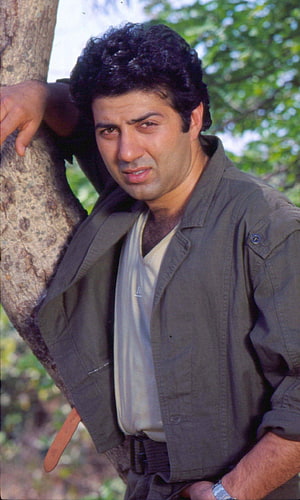 Sunny Deol Leaning Against Tree Trunk in Olive Jacket and White Shirt HD phone wallpaper