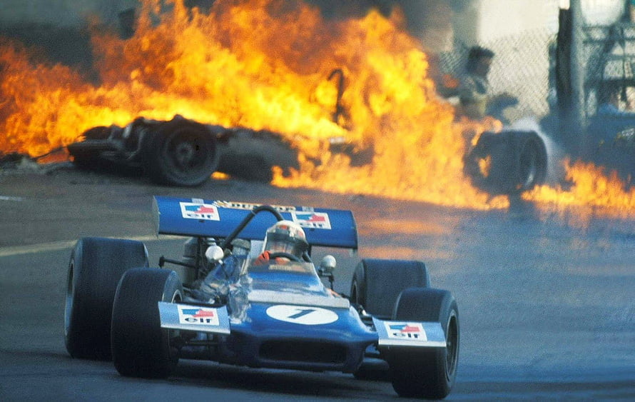 Vintage Formula 1 race car on track with burning wreckage in background HD wallpaper