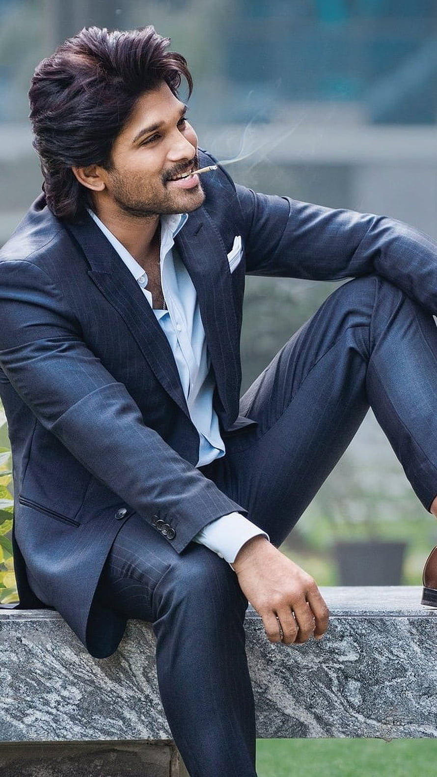 Allu Arjun in a dark suit sitting on a stone ledge with a cigarette HD phone wallpaper