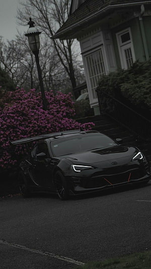 Black Toyota GR86 with rear wing parked on a residential street beside blooming azalea bushes HD phone wallpaper
