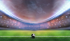 Soccer ball on a green pitch under bright stadium floodlights with a dramatic cloudy sky HD wallpaper