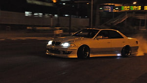 White Nissan Skyline R32 drifting through a city intersection at night with smoke and underglow HD wallpaper