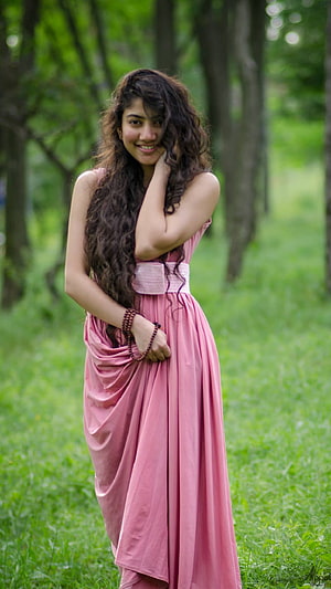 Sai Pallavi in a flowing pink dress standing in a lush green forest clearing HD phone wallpaper