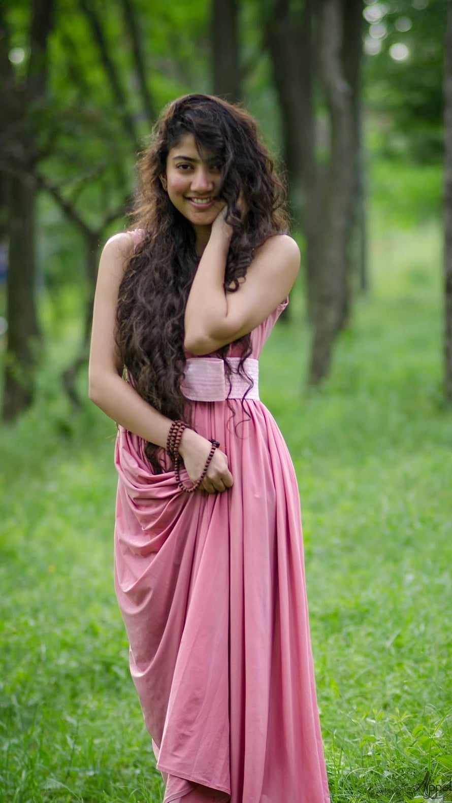 Sai Pallavi in a flowing pink dress standing in a lush green forest clearing HD phone wallpaper