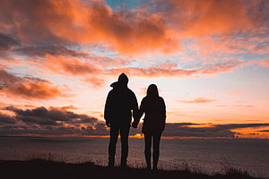 Silhouetted couple holding hands on a coastal cliff at sunset with dramatic orange clouds HD wallpaper