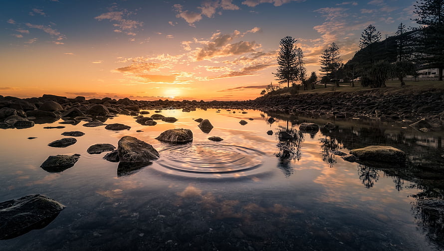Serene Sunset Over Rocky Shallows With Pine Trees And Water Ripples HD wallpaper