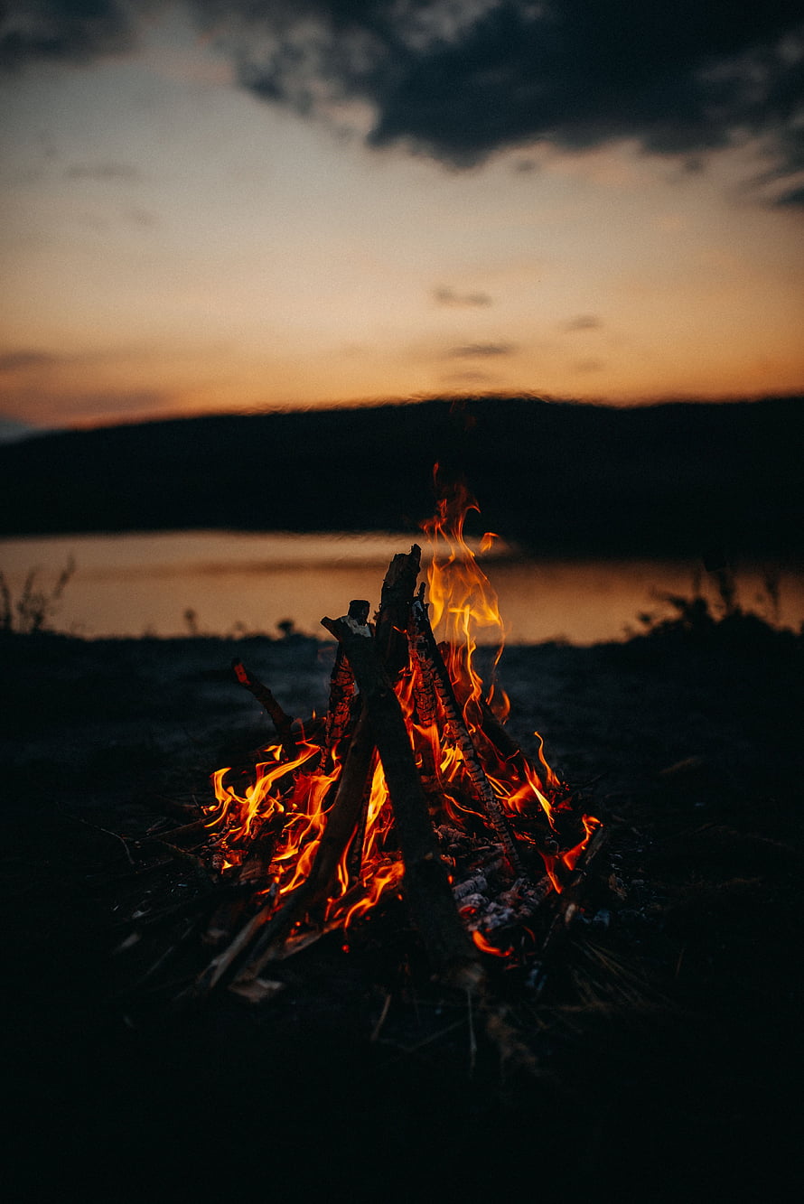 Campfire burning at dusk beside a calm lake with distant hills under a twilight sky HD phone wallpaper