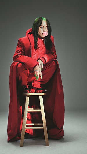 Billie Eilish in oversized red outfit with green hair blowing a pink bubblegum bubble on a wooden stool HD phone wallpaper