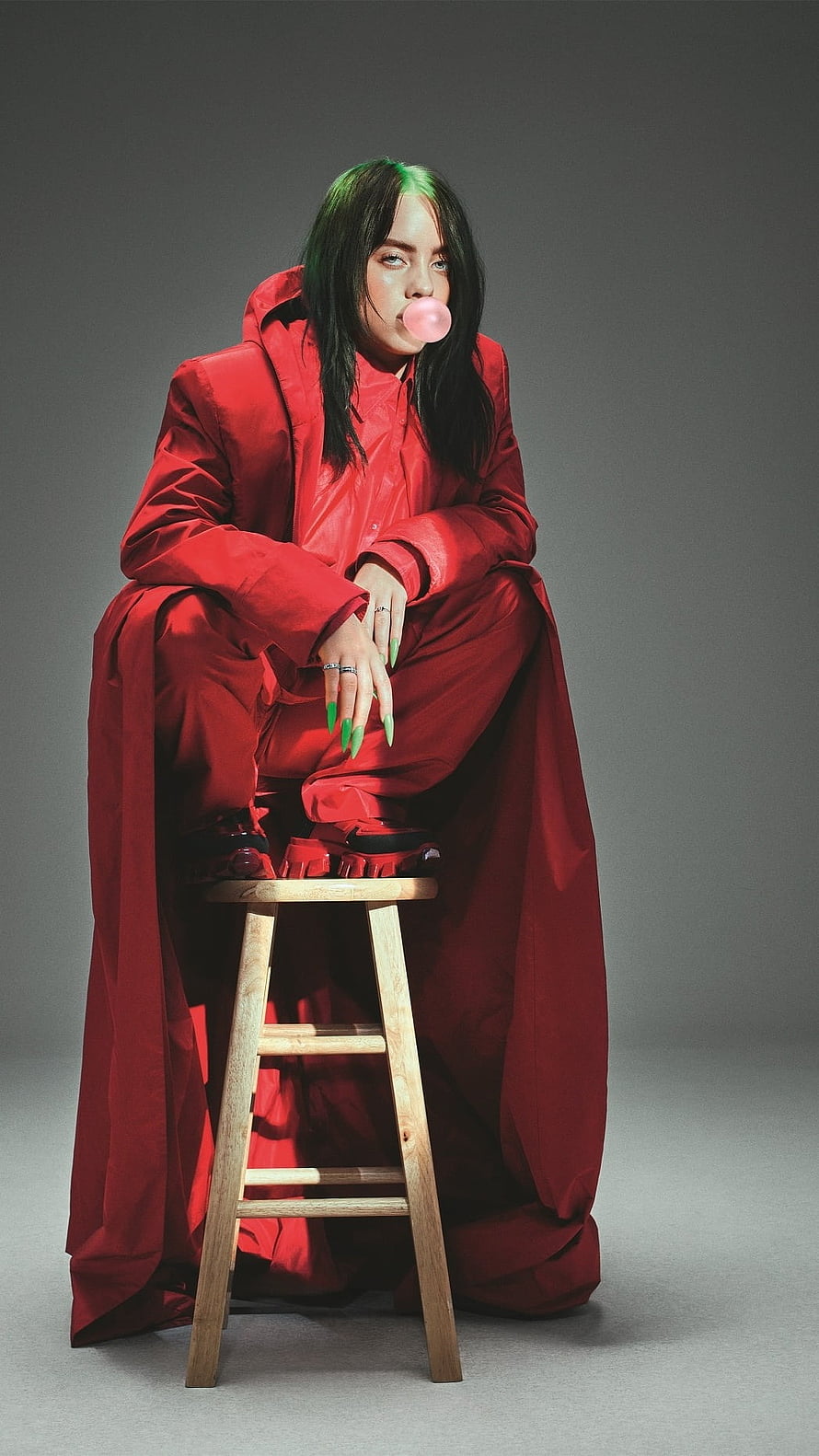 Billie Eilish in oversized red outfit with green hair blowing a pink bubblegum bubble on a wooden stool HD phone wallpaper
