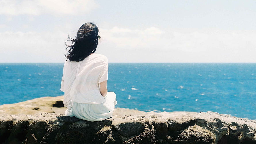 Woman in white dress sitting on rocky cliff overlooking the blue ocean HD wallpaper